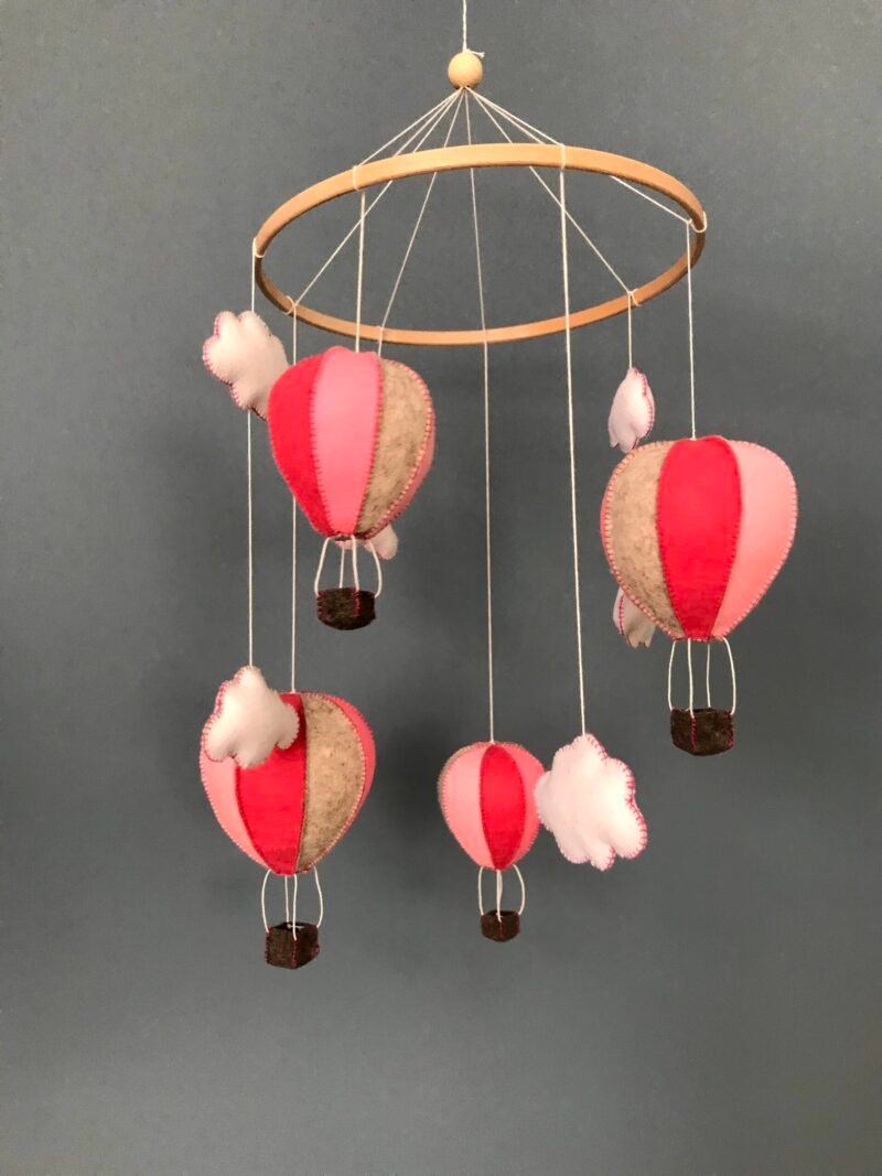 Mobiel voor in de kinderkamer bestaande uit een houten ring met daaraan figuren van wolvilt hangend. Er hangen 4 luchtballonnen in roze tinten en 6 witte wolkjes aan, die met de hand zijn genaaid.