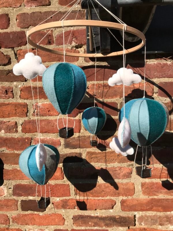 Mobiel van een houten ring met daaraan 4 blauwe luchtballonnen en 6 witte wolkjes van wolvilt. Op de achtergrond een muur van bakstenen