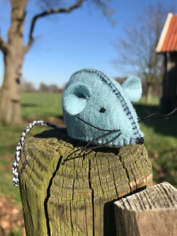 Lichtblauw wolvilt muisje op een paaltje met gras, bomen en blauwe lucht op de achtergrond