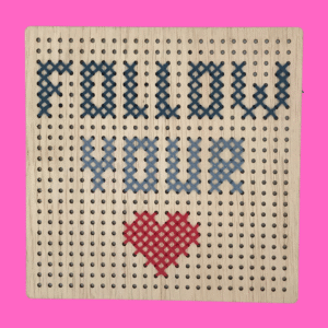 Houten borduurplankje met gaatjes. De tekst Follow your heart is erop geborduurd waarbij het woord heart vervangen is door een tekening van een hartje. De kleuren van het borduursel is donker en licht blauw met een rood hartje.
