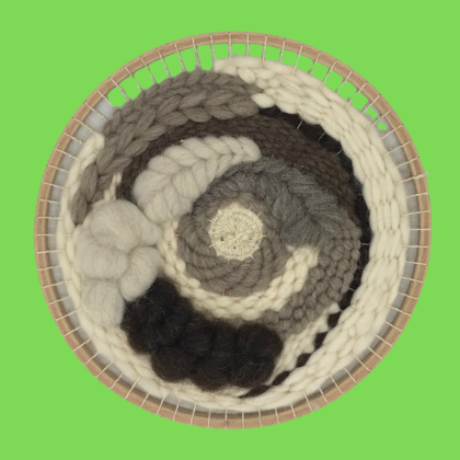 Houten weefcirkel waarop met verschillende soorten echte wol geweven is. De kleuren zijn bruin, beige en ecru en door de verschillende technieken zijn variërende texturen ontstaan. De achtergrond van de foto is fel groen.