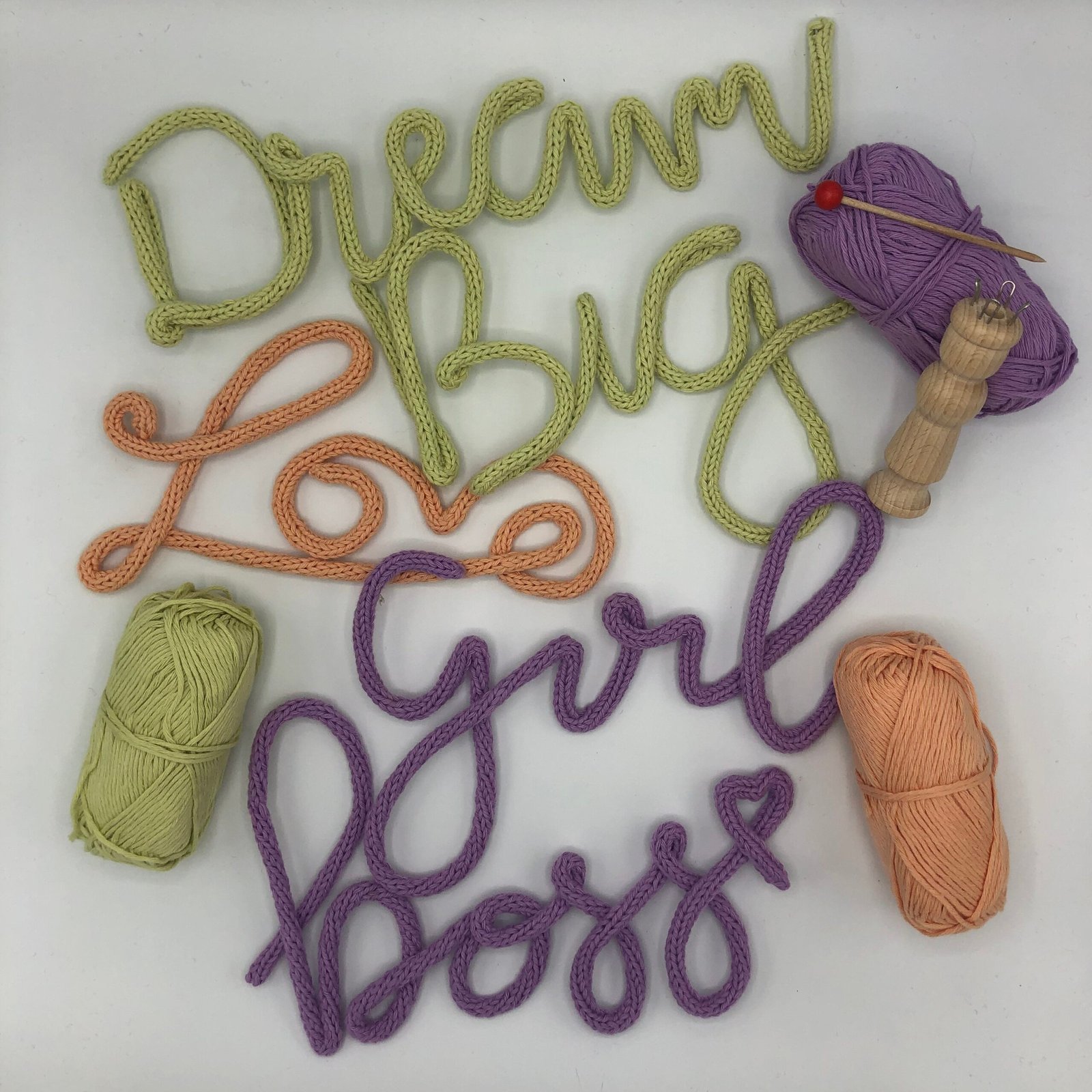 Gepunnikte woorden van katoen in pastelgroen, perzik en paars — Dream Big, Love en Girl Boss — omringd door bolletjes katoen garen en een punnikklosje.